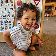 Keanu is registered to the contest to win money with this photo: toddler, child, curly_hair, striped_onesie, smiling, indoor, carpet, educational_posters, alphabet, shapes, playroom, learning, cute, happy, crouching, person, baby, early_learning, colorful, floor