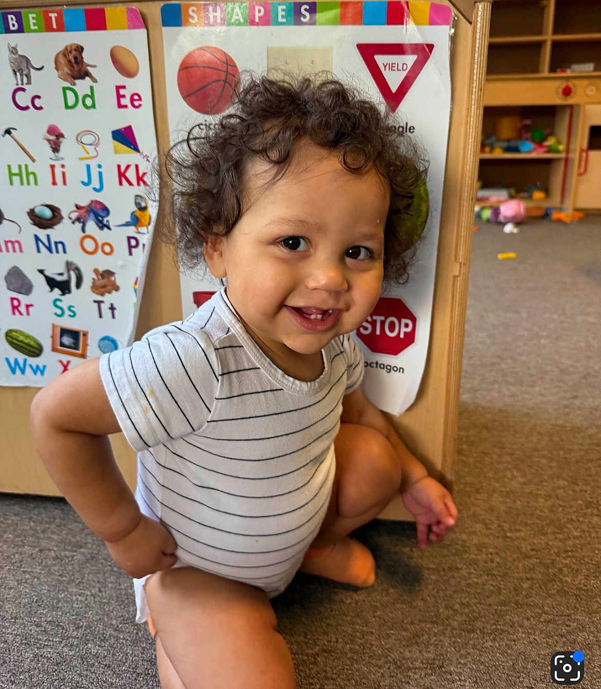 Keanu is registered to the contest to win money with this photo: toddler, child, curly_hair, striped_onesie, smiling, indoor, carpet, educational_posters, alphabet, shapes, playroom, learning, cute, happy, crouching, person, baby, early_learning, colorful, floor
