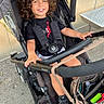Leyann participe au concours pour gagner de l'argent avec cette photo : child, smiling, curly_hair, stroller, sneakers, black_clothing, sidewalk, bench, bag, outdoor, happy, person, seat, casual, daytime, urban, metal, wheels, accessory, young