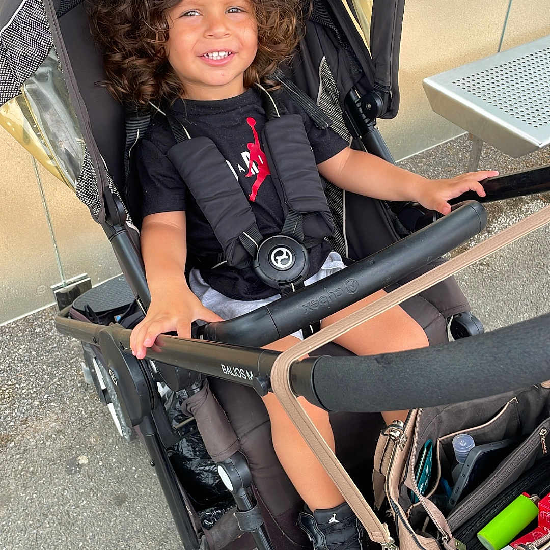 Leyann participe au concours pour gagner de l'argent avec cette photo : child, smiling, curly_hair, stroller, sneakers, black_clothing, sidewalk, bench, bag, outdoor, happy, person, seat, casual, daytime, urban, metal, wheels, accessory, young