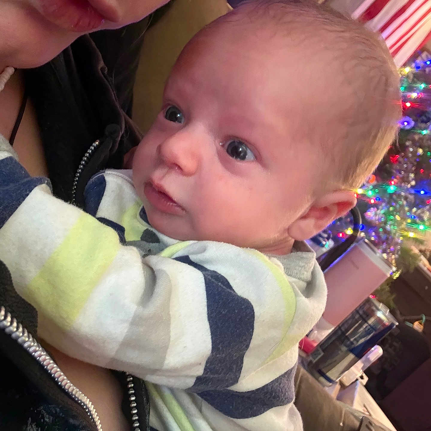 Kody is registered to the contest to win money with this photo: adult, baby, christmas_tree, closeup, clothing, couch, curious, expression, eyes, face, head, holding, holiday_lights, indoor, infant, mouth, nose, person, skin, striped_shirt