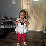child, toddler, indoor, floor, door, shoes, shoe_rack, wall, light_switch, window, glass, clothing, red_pants, white_top, crocs, bracelet, hair, person, standing, gesture