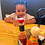 child, toddler, pigtails, pink_shirt, table, wooden_table, hot_sauce, condiment_basket, restaurant, smiling, happy, sitting, food, bottle, spices, bracelet, indoor, cute, person, face