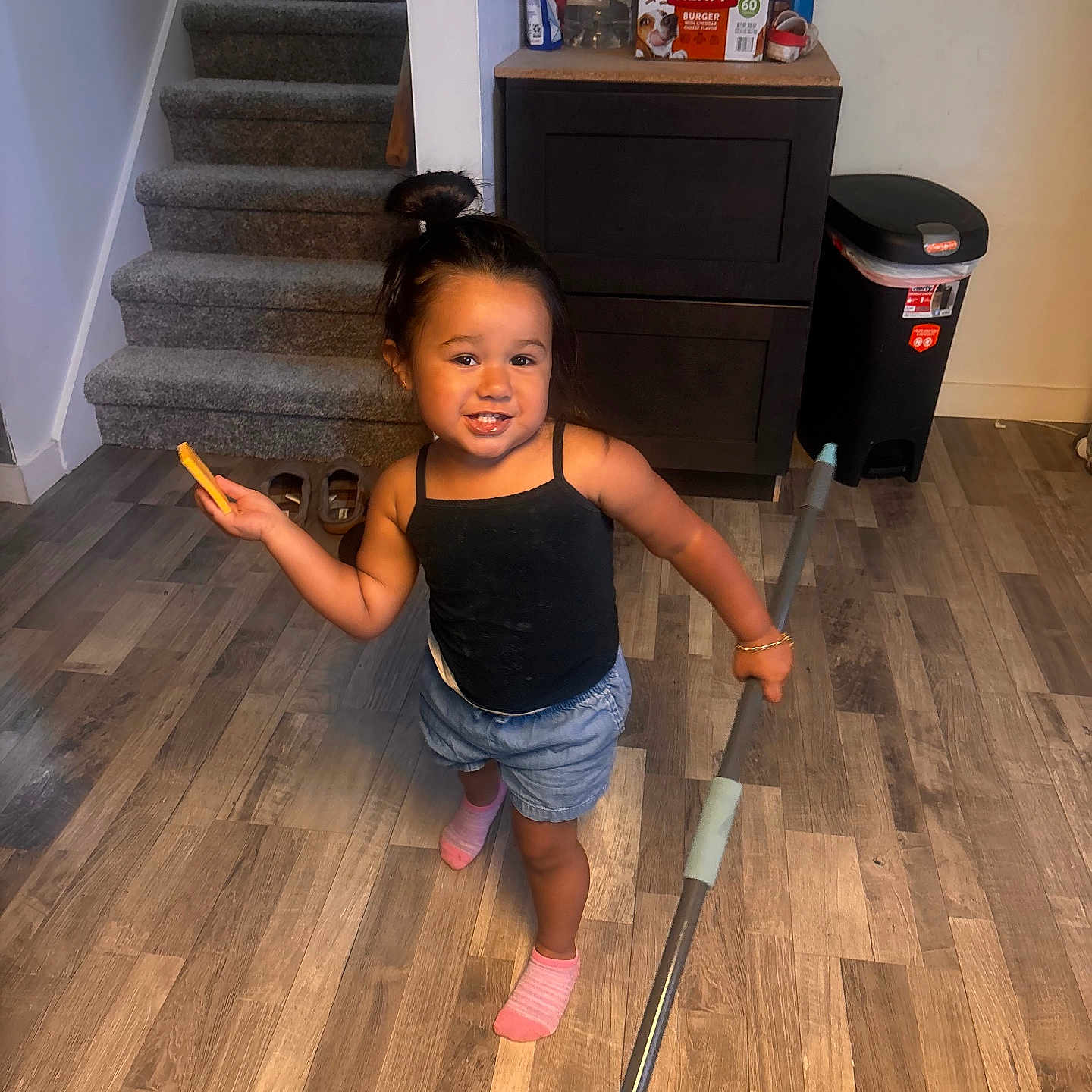 Marisol is registered to the contest to win money with this photo: black_tank_top, bracelet, cabinet, child, cleaning, floor, hair_bun, home, indoor, mop, pink_socks, playful, shorts, smiling, snack, stairs, toddler, toy, trash_can, wooden_floor