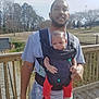 Z'Uriel is registered to the contest to win money with this photo: baby, adult, infant, baby_carrier, front_carrier, wooden_deck, park, trees, fence, railings, sneakers, red_pants, tshirt, beard, outdoor, daylight, portrait, holding, person, family_moment