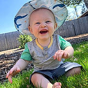 Cooper joined the competition — help win amazing prizes! baby, child, smiling, hat, sunhat, grass, outdoor, blue_sky, fence, tree, greenery, happy, sitting, casual_clothing, shorts, toddler, daylight, nature, portrait, playful