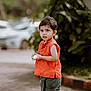 barefoot, casual_clothing, child, curious, cute, daylight, explorer, flower, green_pants, nature, orange_top, outdoor, pavement, plants, portrait, small_person, standing, toddler, urban, young_child