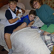 Freya is registered to the contest to win money with this photo: bed, blanket, boy, carpet, casual_clothing, charger, child, curly_hair, dog, friendship, glasses, happy, headboard, indoor, pet, phone, pillow, plaid_blanket, relaxed, smile