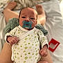 baby, pacifier, onesie, infant, legs, hand, face, person, patterned_clothing, blurred_background, skin, newborn, indoor, resting, cute, small, fist, comfort, soft, lying