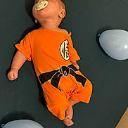 Bruno a rejoint le concours — aidez-le/la à gagner de superbes lots ! baby, baby_clothing, balloon, blue_balloon, celebration, child, curious_look, cute, dark_background, decor, indoors, infant, lying_down, orange_outfit, pacifier, playful, resting, small_person, soft_lighting, yellow_balloon