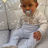 Noé participe au concours pour gagner de l'argent avec cette photo : baby, pacifier, pajamas, couch, white, plastic_cover, child, indoor, seated, cute, soft_clothing, face, hand, footwear, expression, furniture, home, carrot, bear_illustration, curious