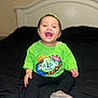 Genesis is registered to the contest to win money with this photo: toddler, child, smiling, happy, indoor, bed, clothing, green_shirt, sitting, face, person, feet, shoes, black_pants, head, hair, expression, joy, infant, portrait