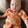baby, child, pumpkin_shirt, orange_shorts, cushion, sofa, blue_eyes, curly_hair, casual_clothing, indoor, relaxed, portrait, cute, festive, halloween_theme, infant, comfortable, soft_lighting, closeup, person