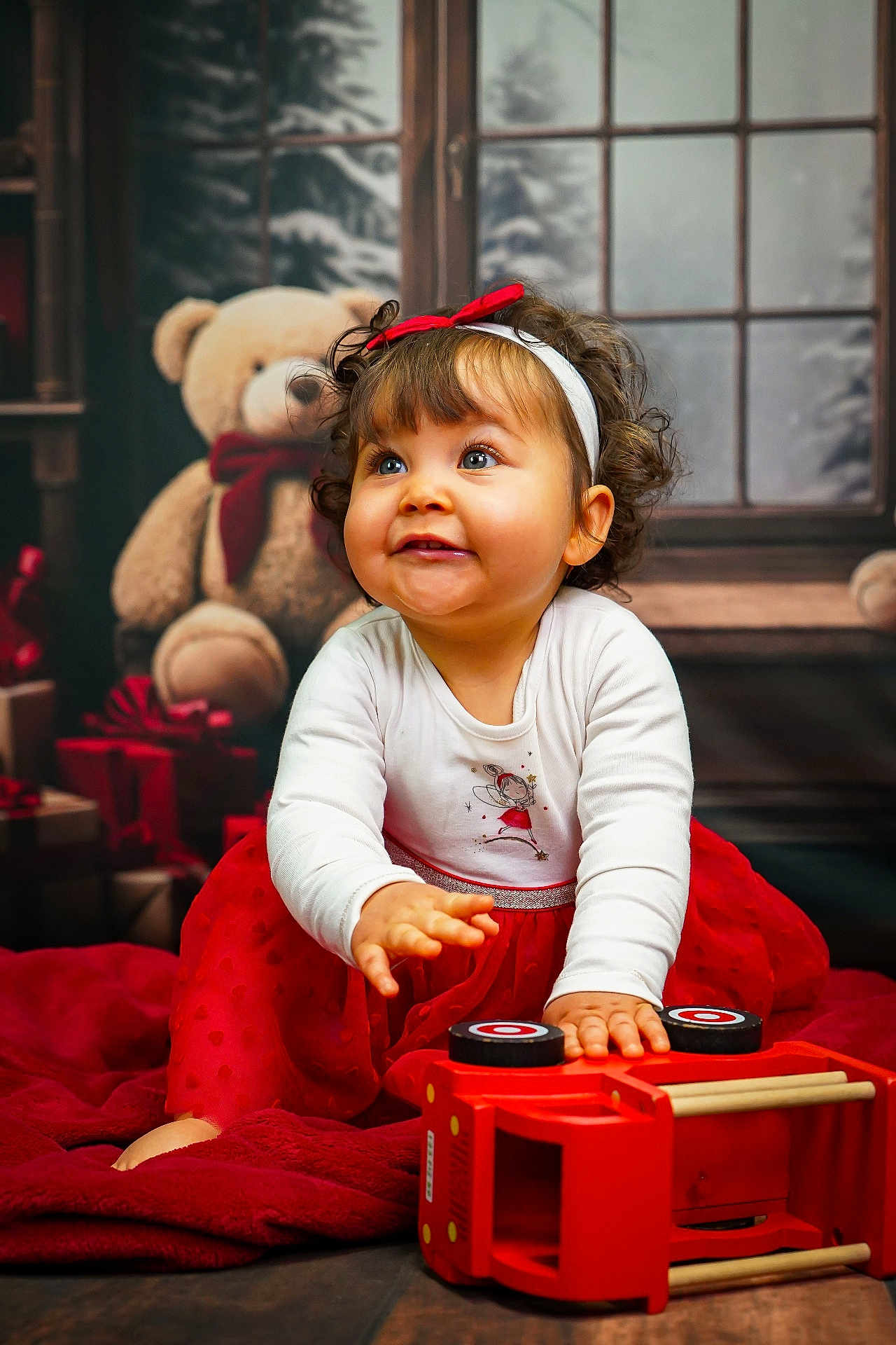 Mya participe au concours pour gagner de l'argent avec cette photo : toddler, child, curly_hair, red_bow, white_shirt, red_skirt, toy, wooden_toy, teddy_bear, gift_boxes, blanket, indoor, window, floor, happy, smiling, playful, holiday, festive, portrait