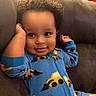 baby, child, infant, smiling, portrait, couch, indoor, seated, blue_outfit, patterned_clothing, mustache_pattern, pacifier, curly_hair, brown_skin, face, eyes, hands, warm_lighting, cozy, closeup