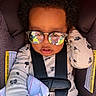 baby, child, infant, car_seat, sunglasses, portrait, sitting, harness, buckled, curly_hair, onesie, blanket, fashion, reflection, indoors, vehicle, close_up, face, safe, adorable