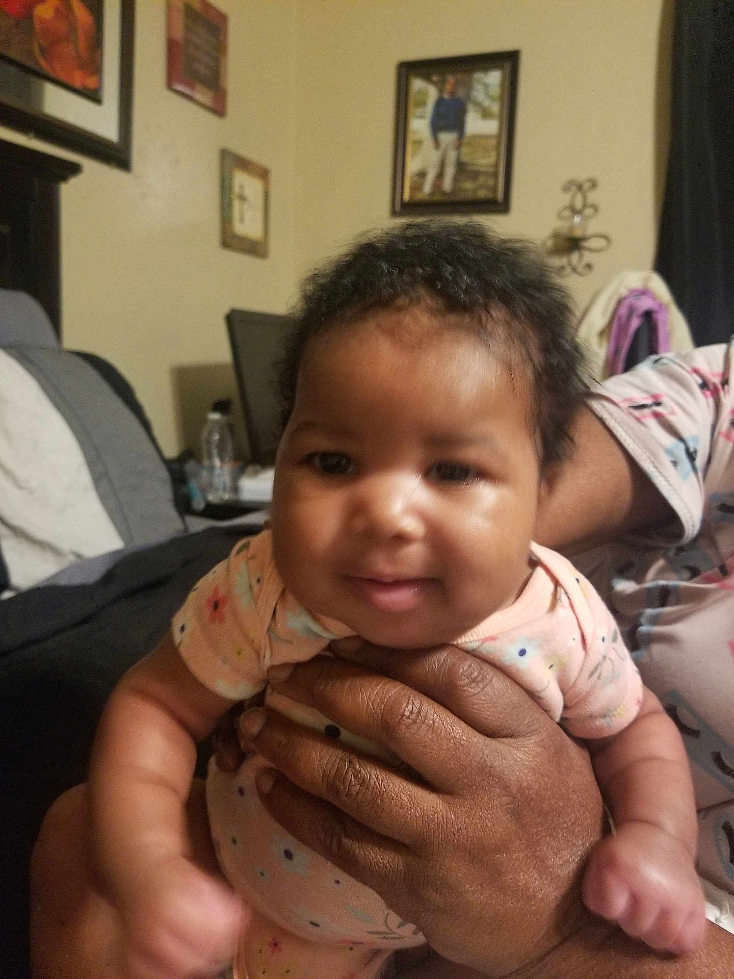 De'Nai joined the competition — help win amazing prizes! baby, infant, hands, person, indoor, onesie, smile, face, curly_hair, furniture, bed, picture_frame, wall_art, decor, adult_hand, child, cozy, home, portrait, holding