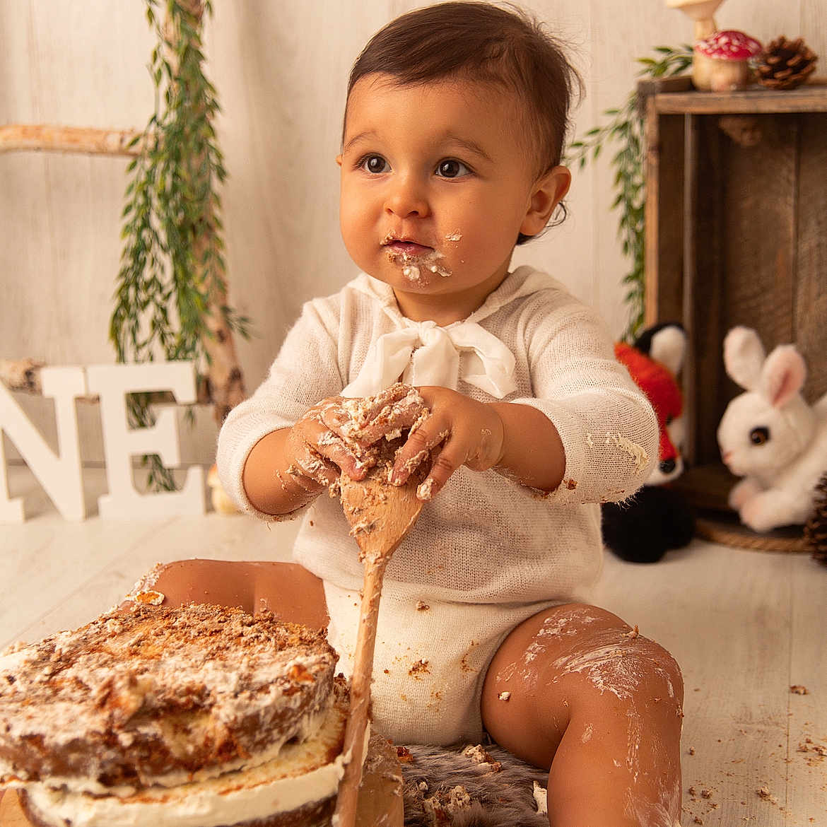 Noa participe au concours pour gagner de l'argent avec cette photo : baby, birthday, cake, celebration, child, cute, decor, face, frosting, greenery, hands, indoor, legs, messy, plush_toy, sitting, smile, white_clothing, wooden_floor, wooden_spoon