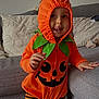 Noa a rejoint le concours — aidez-le/la à gagner de superbes lots ! child, toddler, pumpkin_costume, orange, green, striped_pants, striped_leggings, cute, smiling, tongue_out, indoor, living_room, sofa, pillows, costume, playful, baby, person, holiday, halloween