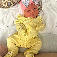 Magnolia is registered to the contest to win money with this photo: newborn, baby, infant, child, portrait, lying_down, yellow_outfit, bee_pattern, striped_hat, pink_hat, duck_socks, bed, blanket, pillow, cushion, indoor, close_up, face, serious_expression, cozy