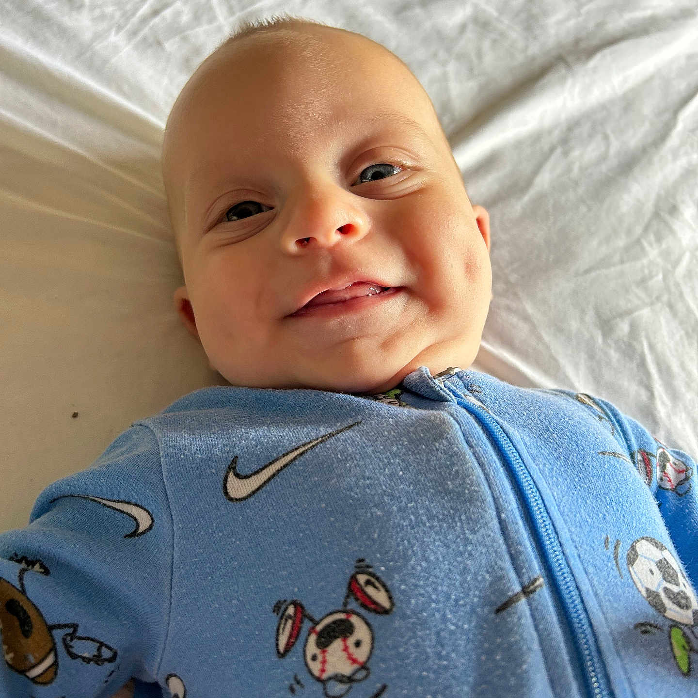 Oakley is registered to the contest to win money with this photo: baby, bed, blue_clothing, child, cute, eyes, face, happy, head, indoor, infant, lying_down, mouth, onesie, portrait, sheet, skin, smile, soft_light, sports_print