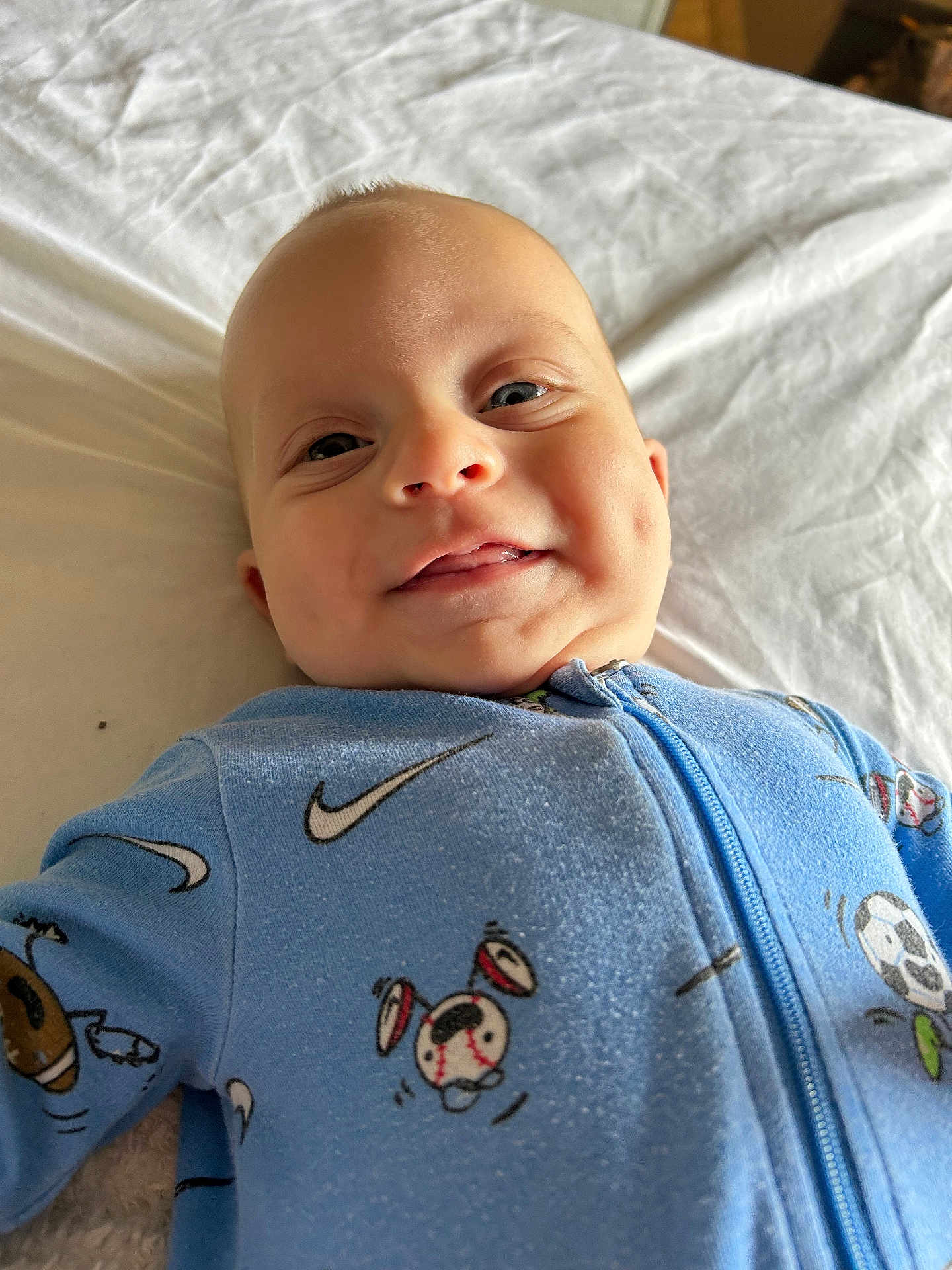 Oakley is registered to the contest to win money with this photo: baby, child, infant, smile, face, onesie, blue_clothing, sports_print, lying_down, bed, sheet, happy, cute, portrait, skin, head, eyes, mouth, indoor, soft_light