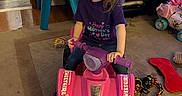 Brooklyn is registered to the contest to win money with this photo: child, girl, toddler, indoor, living_room, toy_vehicle, quad_bike, pink, sitting, riding, long_hair, purple_shirt, smiling, television, cartoon, toys, playroom, carpet, home, portrait