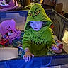 child, toddler, hooded_costume, green_costume, monster_costume, playpen, toy, pink_chair, stuffed_animal, indoors, home, playroom, sitting, standing, face, portrait, curious, colorful, baby_gear, household_clutter