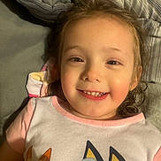 Brooklyn is registered to the contest to win money with this photo: child, girl, smiling, portrait, face, indoors, bedroom, bed, pillows, bedding, lying_down, pink_shirt, cartoon, cat_design, brown_hair, happy, close_up, cozy, pointing, photograph