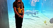Isabella is registered to the contest to win money with this photo: child, girl, aquarium, shark, underwater, marine_life, fish, portrait, standing, indoors, blue_lighting, glass_tank, aquatic, exhibit, pikaachu_shirt, long_hair, smiling, travel, photography, family_outing