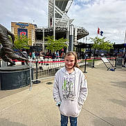 Isabella is registered to the contest to win money with this photo: child, girl, stadium, sports_venue, outdoor, portrait, standing, hoodie, jeans, sneakers, statue, crowd, flags, architecture, entrance, barriers, concrete, cloudy_sky, travel, daylight