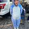 Isabella is registered to the contest to win money with this photo: child, dinosaur_onesie, blue, hood, costume, standing, portrait, smiling, face, outdoor, parking_lot, cars, suv, folding_chair, stool, sneakers, autumn, daylight, playful, photograph