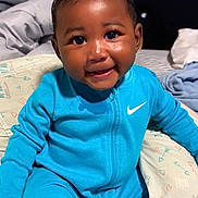 Kyeir joined the competition — help win amazing prizes! baby, child, smile, blue_clothing, nike_logo, onesie, bed, bedding, pillow, indoor, portrait, happy, cute, sitting, infant, young_child, face, skin, person, cozy