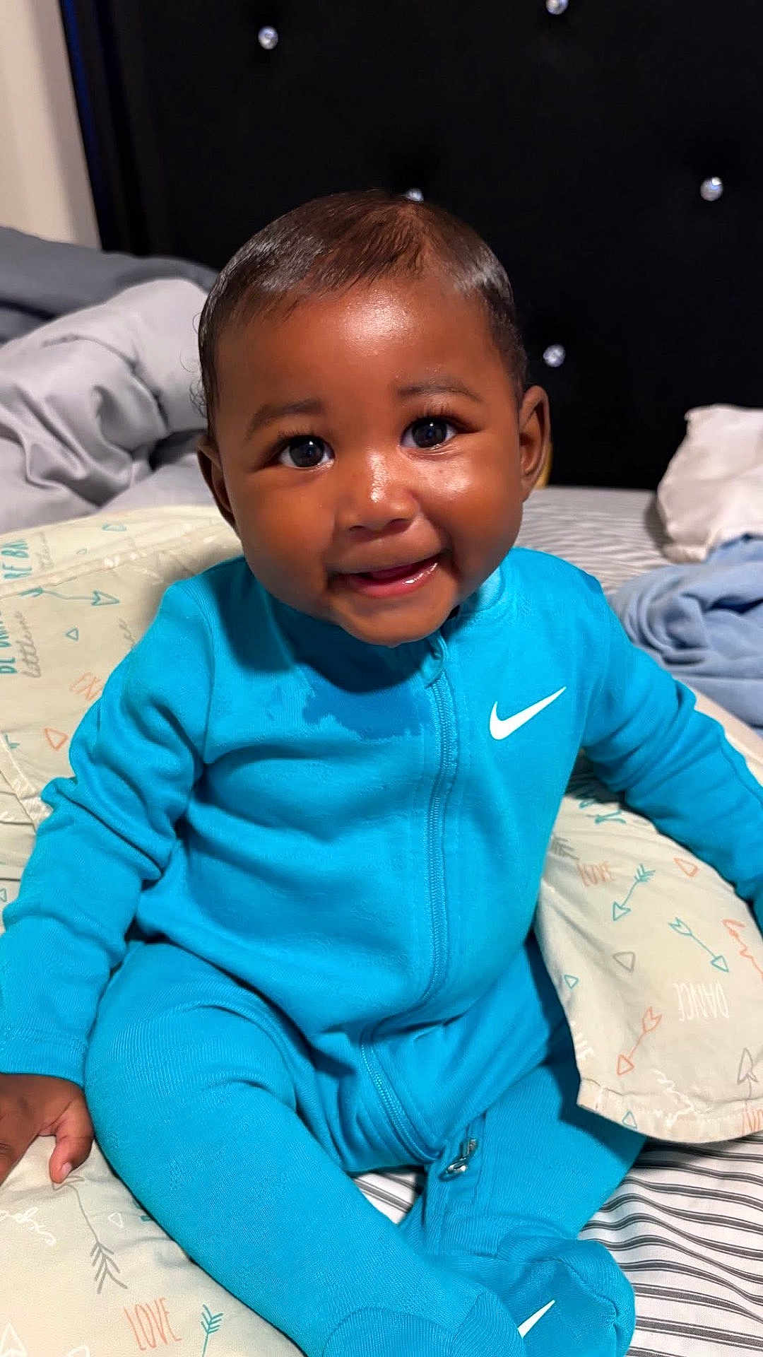 Kyeir joined the competition — help win amazing prizes! baby, child, smile, blue_clothing, nike_logo, onesie, bed, bedding, pillow, indoor, portrait, happy, cute, sitting, infant, young_child, face, skin, person, cozy