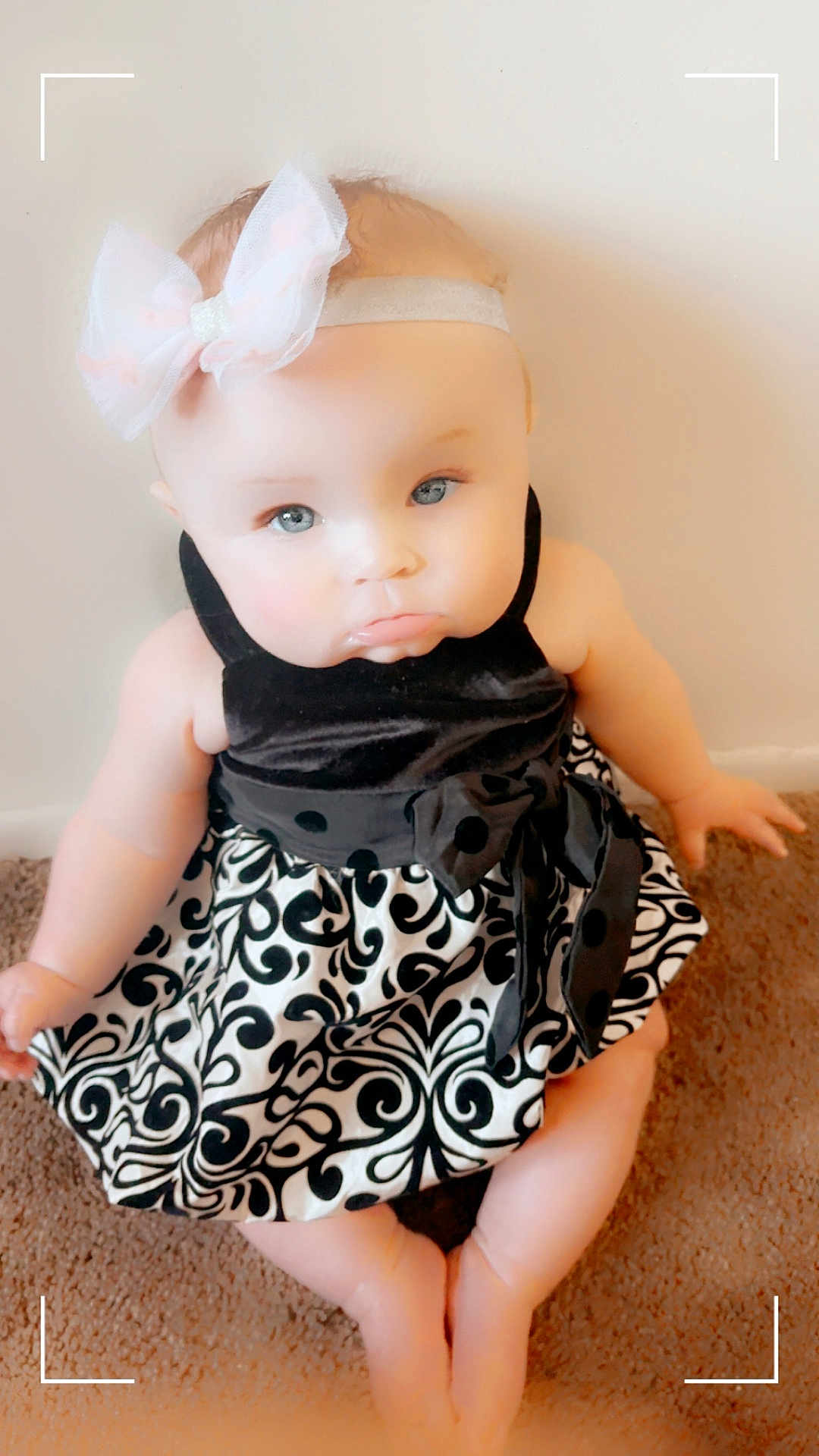 Isabella is registered to the contest to win money with this photo: baby, child, dress, headband, bow, carpet, pout, cute, sitting, indoor, infant, person, patterned_clothing, black_and_white, soft_skin, happy, small_hands, small_feet, portrait, floor