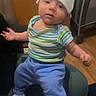 baby, infant, hat, striped_shirt, blue_pants, white_socks, cushion, indoor, wooden_floor, playpen, furniture, child, person, seated, curious, young_child, casual_clothing, home, portrait, cute