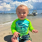 Bobby is registered to the contest to win money with this photo: toddler, child, beach, sand, water, boat, sky, clouds, swim_shirt, swim_trunks, sunny, outdoor, smiling, happy, summer, vacation, nature, shore, sitting, playful