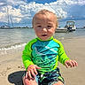toddler, child, beach, sand, water, boat, sky, clouds, swim_shirt, swim_trunks, sunny, outdoor, smiling, happy, summer, vacation, nature, shore, sitting, playful