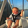 Bobby is registered to the contest to win money with this photo: baby, child, smile, swing, boat, water, sunlight, outdoor, happy, seat, rope, blue_sky, metal, face, person, summer, vacation, cute, safety_harness, legs