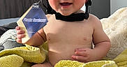 Bobby joined the competition — help win amazing prizes! baby, helmet, towel, indoor, child, holding, curious, joyful, skin, face, person, young_child, furniture, bedroom, blanket, hand, foot, sitting, headgear, toy