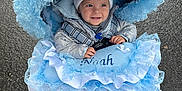Noah is registered to the contest to win money with this photo: baby, child, stroller, blue, white, lace, ribbons, hat, smile, face, clothing, outdoor, pavement, hands, warm_clothing, cute, person, infant, cold_weather, accessory