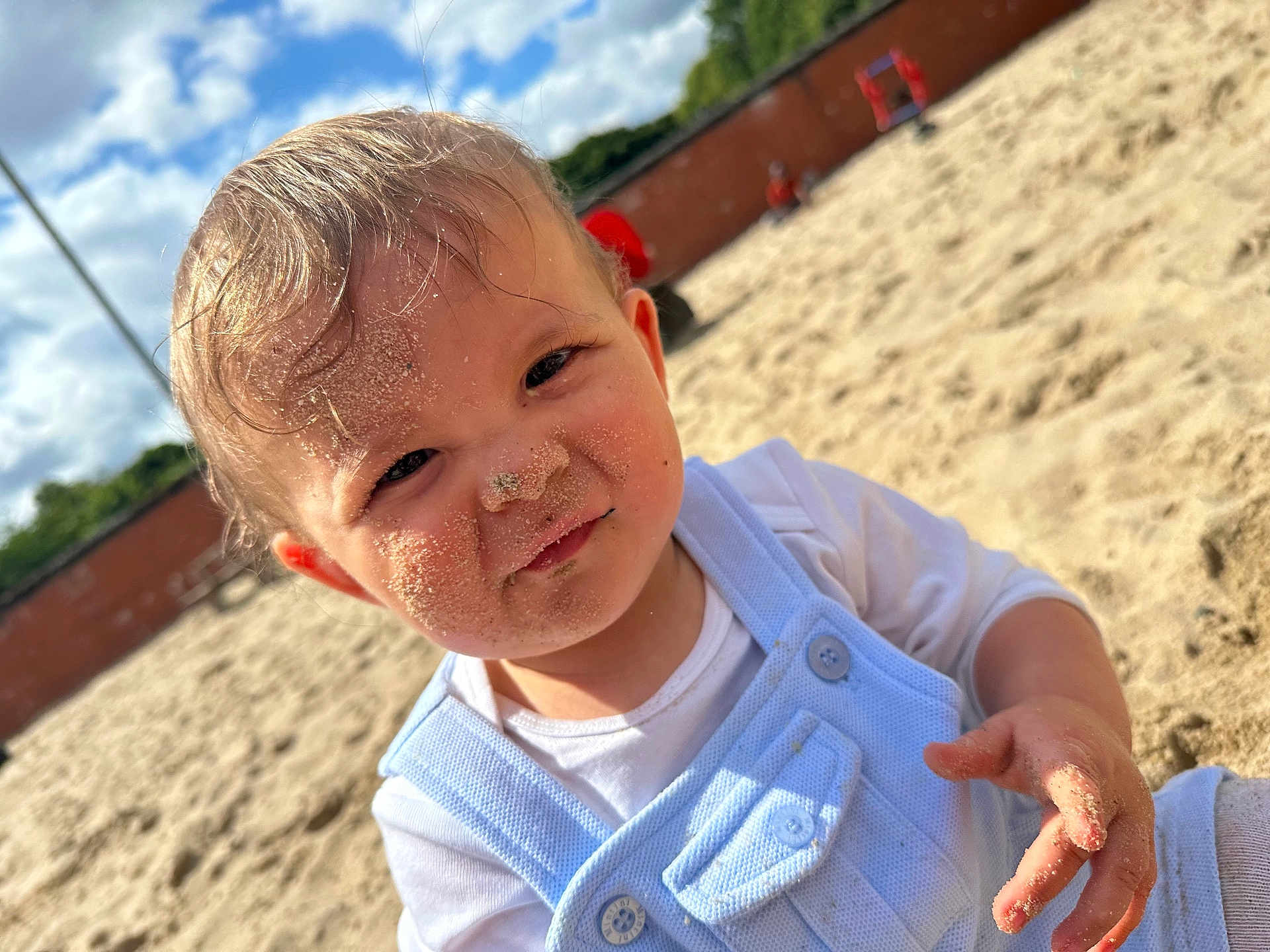 Noah is registered to the contest to win money with this photo: toddler, child, sand, sandbox, outdoor, daylight, blue_sky, clouds, playground, clothing, face, hand, curious, sandy_face, short_hair, white_shirt, overalls, sunlight, smile, young_child
