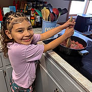 Skye joined the competition — help win amazing prizes! child, smiling, kitchen, cooking, stove, pan, ground_meat, utensils, spices, window, countertop, cabinet, appliance, braided_hair, purple_shirt, happy, indoor, person, food_preparation, home