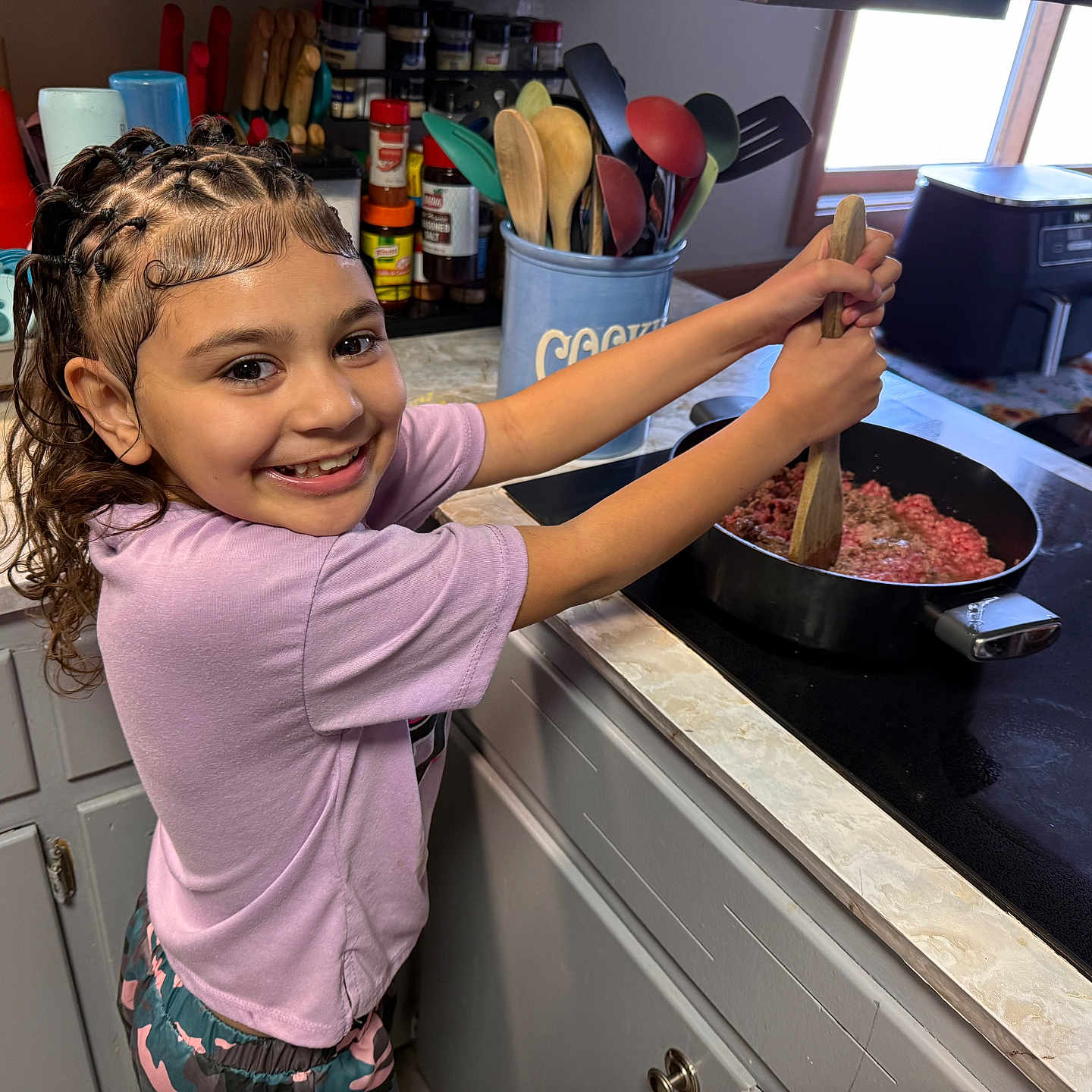 Skye joined the competition — help win amazing prizes! child, smiling, kitchen, cooking, stove, pan, ground_meat, utensils, spices, window, countertop, cabinet, appliance, braided_hair, purple_shirt, happy, indoor, person, food_preparation, home