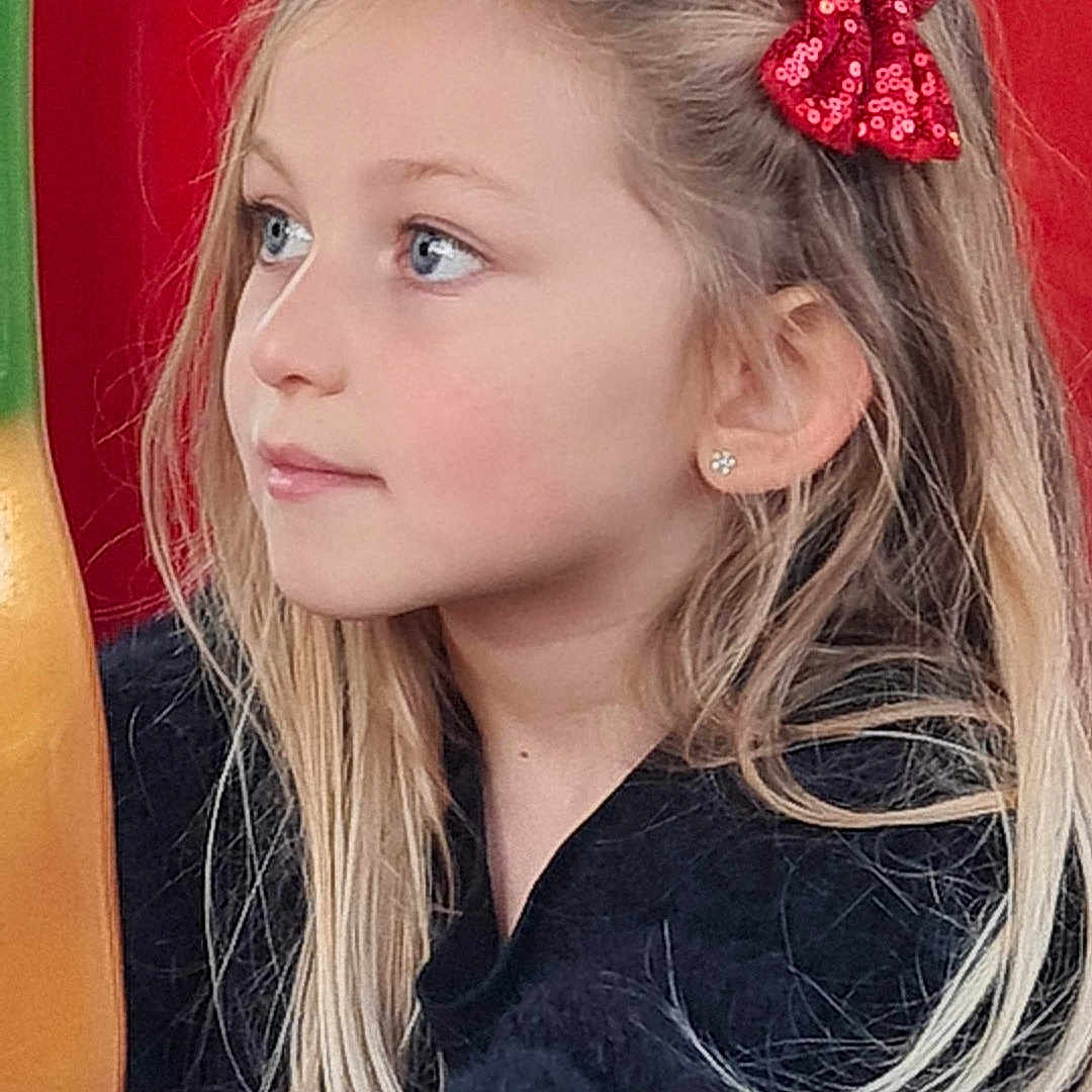 Joy participe au concours pour gagner de l'argent avec cette photo : black_sweater, blonde_hair, blue_eyes, casual_clothing, child, colorful_background, cute, daylight, earrings, girl, innocent, long_hair, outdoor, playground, portrait, red_bow, side_profile, smiling, thoughtful, young