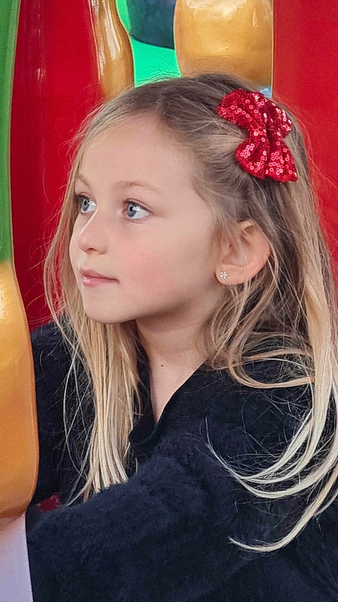 Joy participe au concours pour gagner de l'argent avec cette photo : child, girl, blonde_hair, red_bow, earrings, side_profile, blue_eyes, black_sweater, playground, colorful_background, portrait, young, cute, casual_clothing, long_hair, outdoor, daylight, innocent, thoughtful, smiling
