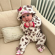 Heidi is registered to the contest to win money with this photo: baby, cow_costume, onesie, hood, footwear, couch, cushion, radiator, indoor, child, cute, plush, seated, clothing, infant, hands, expression, face, adorable, soft