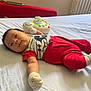 baby, infant, bed, toy, mittens, socks, red_pants, striped_shirt, cartoon_character, white_bed_sheet, couch, radiator, indoors, child, cute, lying_down, playful, home, soft_lighting, curious