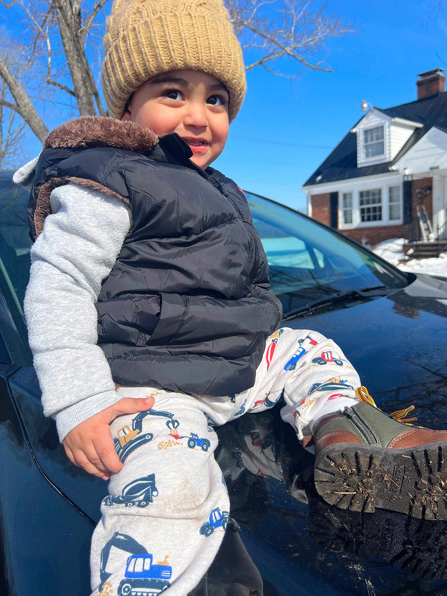 Gabreil is registered to the contest to win money with this photo: toddler, child, hat, vest, car, outdoor, winter, blue_sky, house, smile, pants, boots, tree, sunlight, face, clothing, vehicle, reflection, happy, person