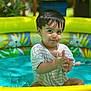 toddler, child, pool, water, inflatable_pool, summer, wet_hair, striped_shirt, outdoor, sunlight, playful, happy, clapping, sitting, colorful, greenery, toys, closeup, baby, fun